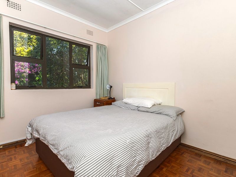 A bedroom featuring parquet flooring, neutral wall tones, and an aluminium-framed window overlooking garden greenery.