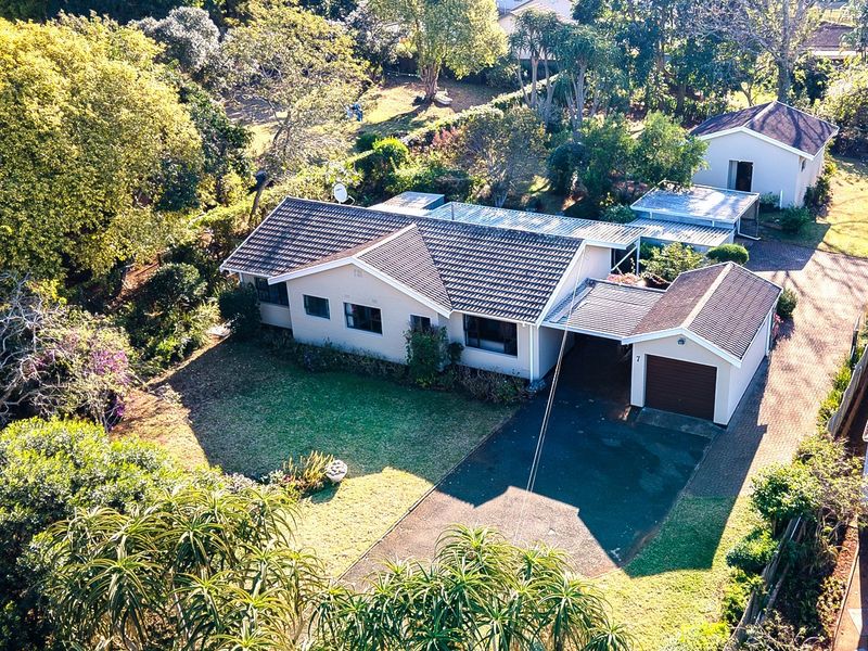 Aerial view of a residential property featuring a main house, detached outbuilding, paved driveway, carport, and established garden surroundings.