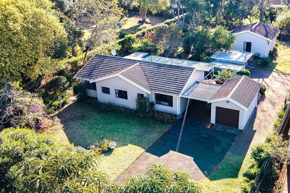 Aerial view of a residential property featuring a main house, detached outbuilding, paved driveway, carport, and established garden surroundings.