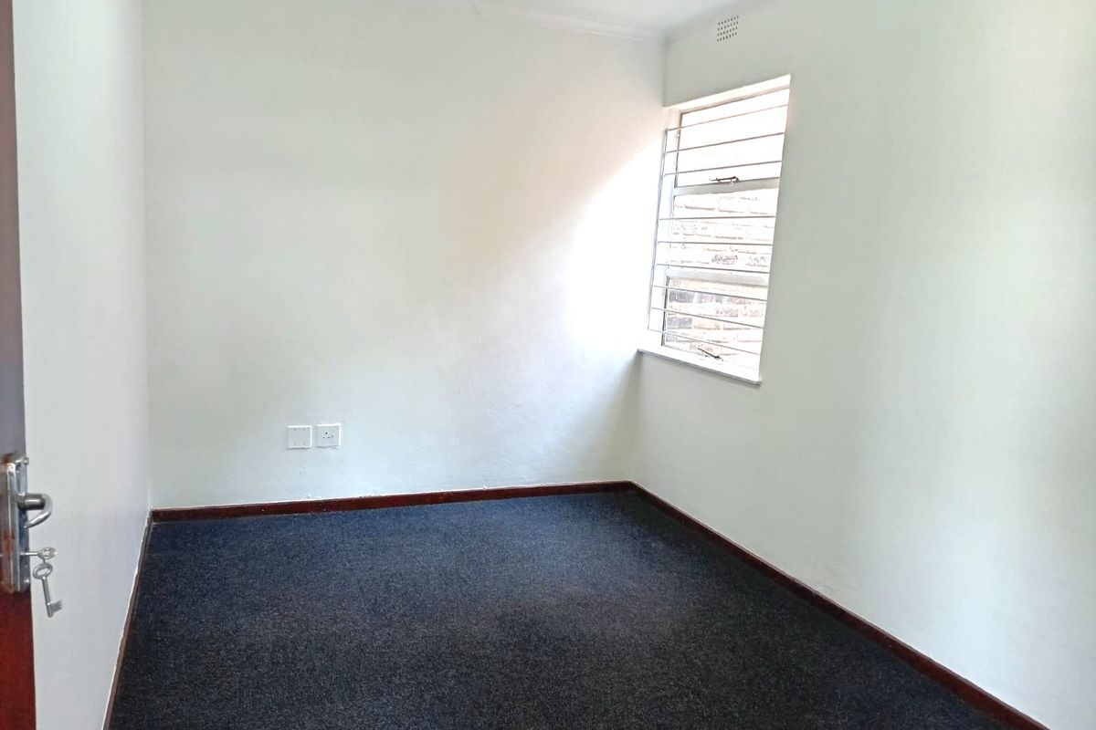 A clean, empty bedroom featuring dark carpeting, white walls, and a louvered window with security bars.