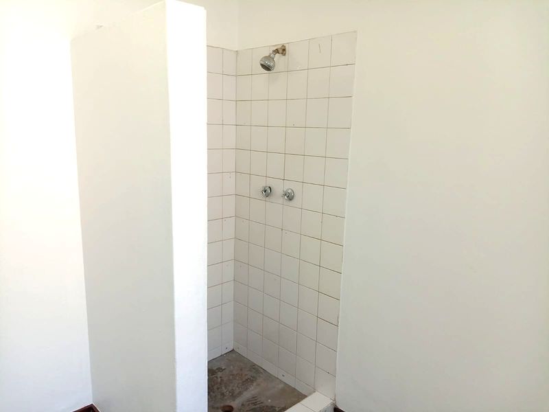 A basic shower enclosure featuring white wall tiles and exposed plumbing fixtures, currently requiring floor finishing and general maintenance.
