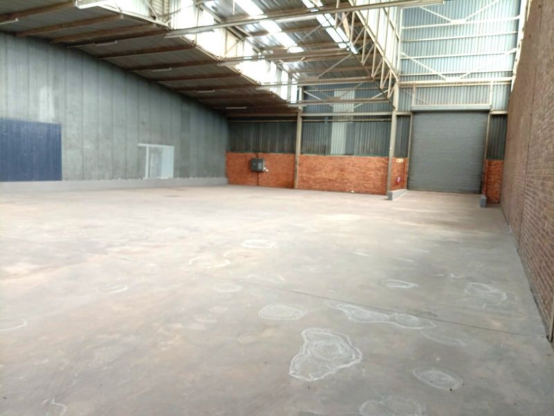 Spacious industrial warehouse unit featuring high ceilings with exposed steel trusses, skylights, concrete flooring, and a large roller shutter door for easy access.
