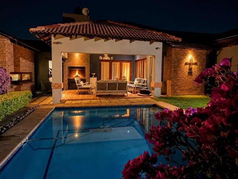 A beautifully lit outdoor entertainment area featuring a swimming pool, covered patio with a built-in fireplace, and classic brick architecture.