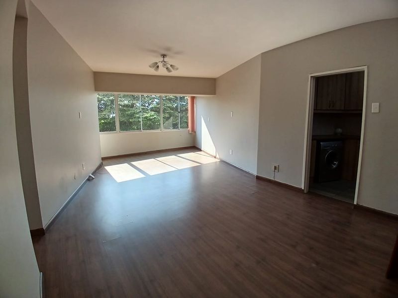 A spacious, empty living room featuring laminate flooring, neutral walls, and a large window providing natural light, with an adjacent utility area.