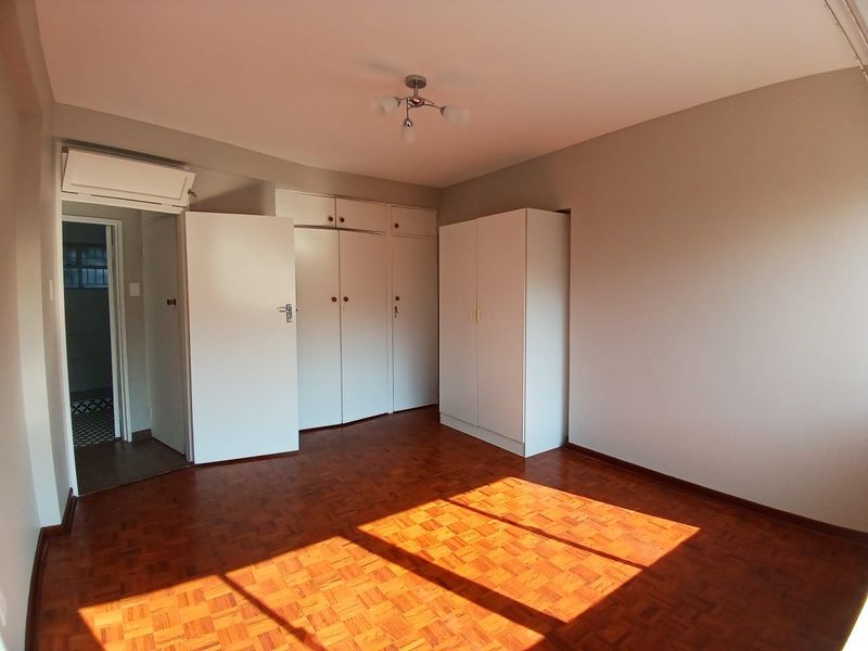 A bright, empty bedroom featuring classic parquet flooring, ample built-in cupboard storage, and an installed air conditioning unit.