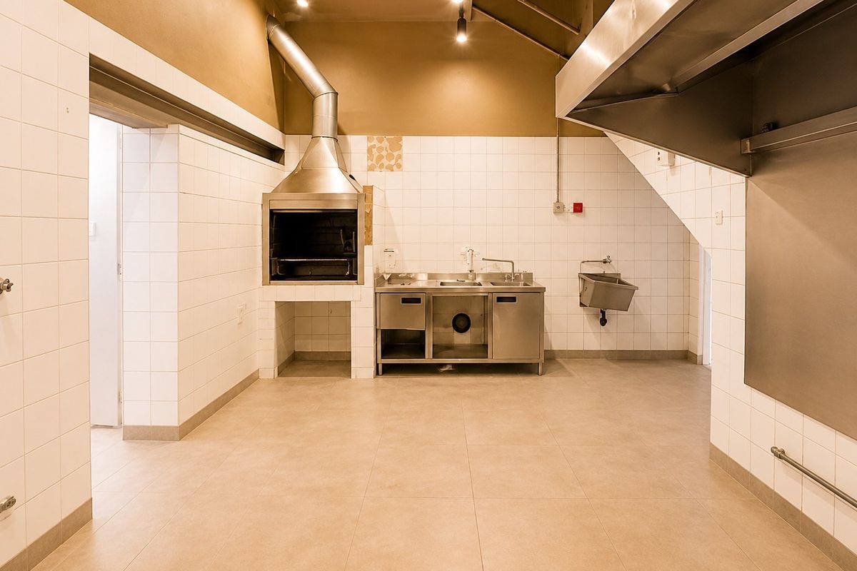 A clean, commercial-grade kitchen area featuring stainless steel fixtures, a built-in braai with extractor, and durable tiled finishes throughout.
