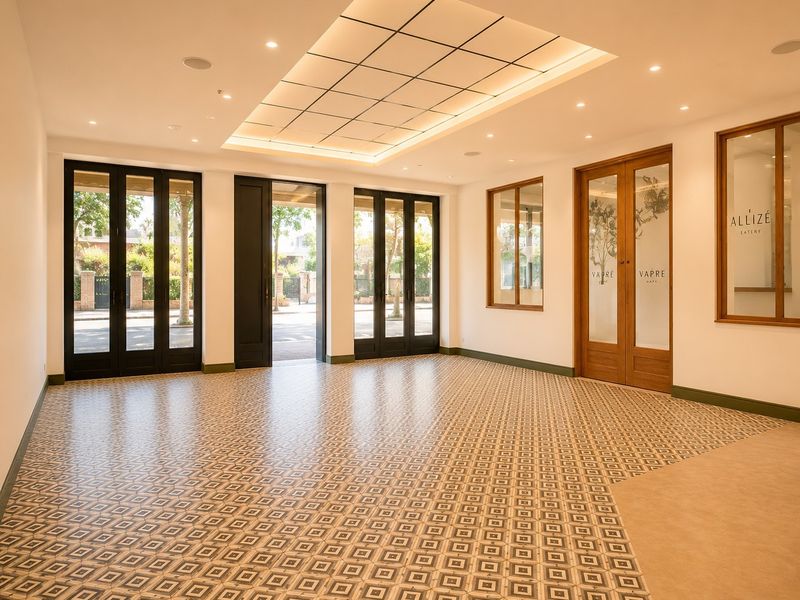 A bright, vacant commercial open-plan space featuring elegant patterned floor tiles, recessed ceiling lighting, and large glass-paneled doors providing natural light and street access.