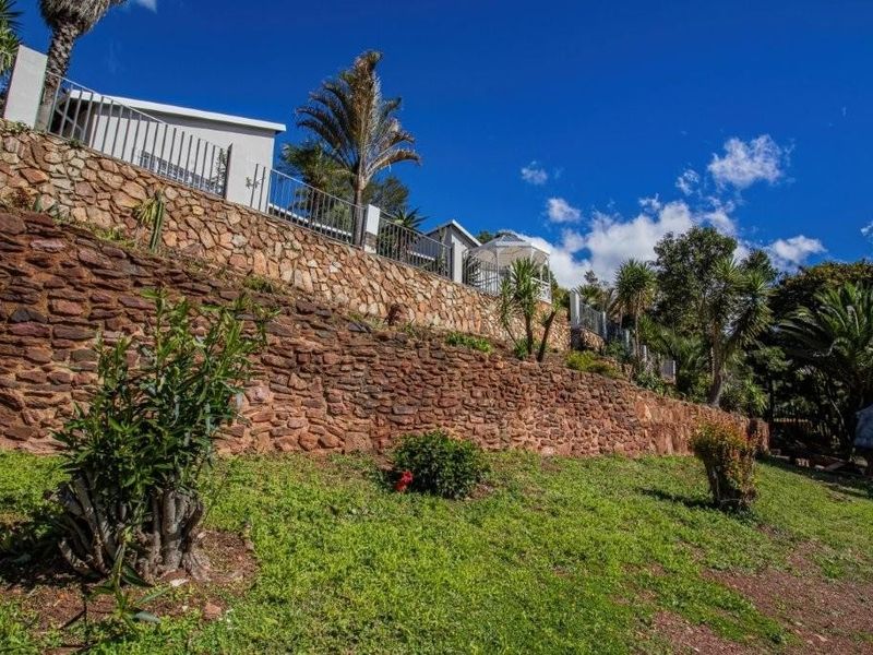 A view of the property's rear garden featuring tiered stone retaining walls, established landscaping, and secure metal perimeter fencing.