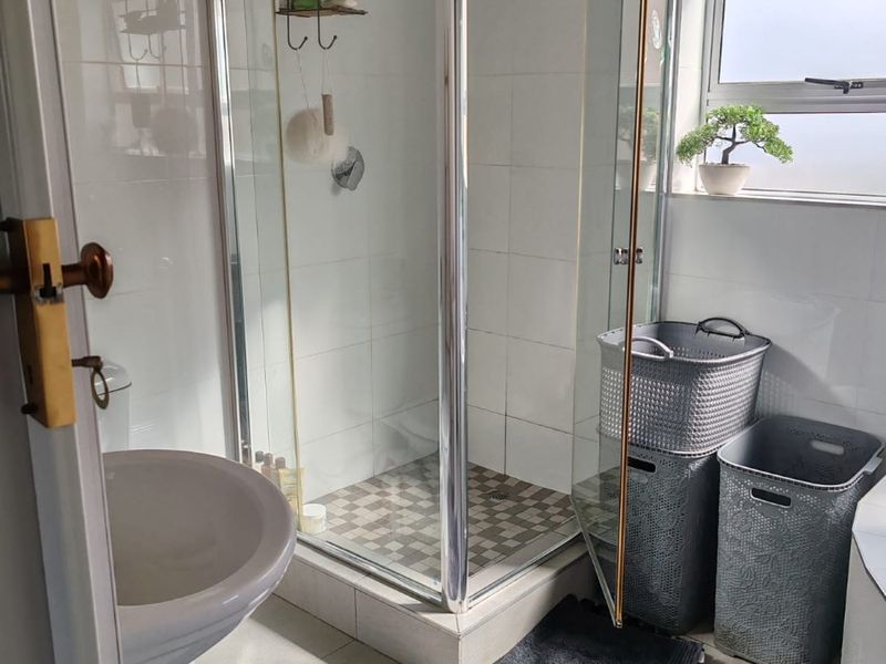 A functional bathroom featuring a pedestal basin, a glass-enclosed shower, and neutral wall and floor tiling with natural light from a frosted window.