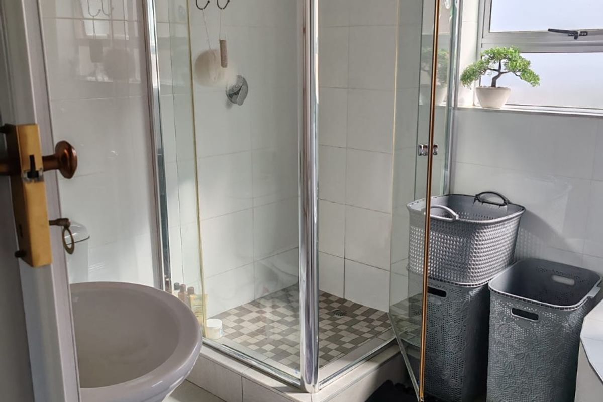 A functional bathroom featuring a pedestal basin, a glass-enclosed shower, and neutral wall and floor tiling with natural light from a frosted window.