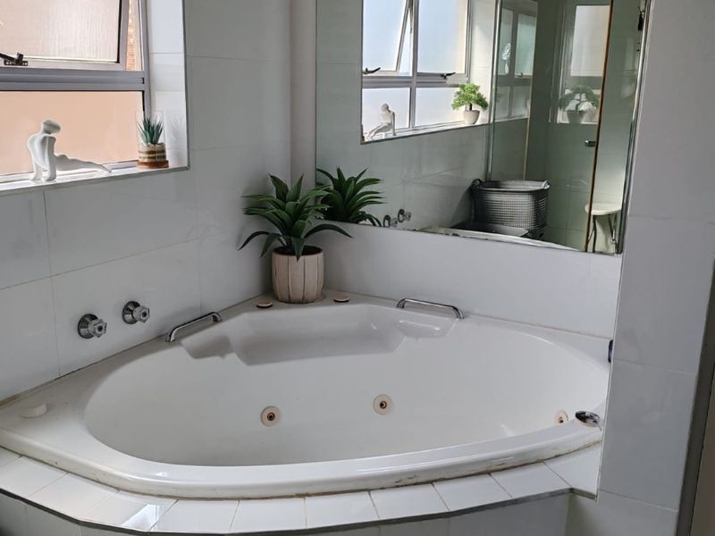 A bathroom featuring a corner bathtub with chrome fixtures, a large wall-mounted mirror, and tiled finishes, offering a functional space with natural light.