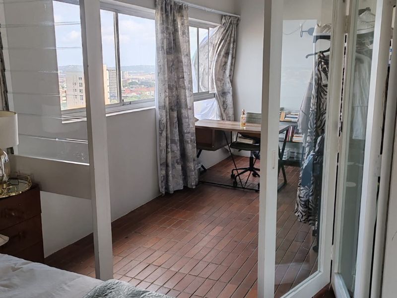 A sunlit home office space featuring terracotta tile flooring, large sliding windows with city views, and access via classic white french doors.
