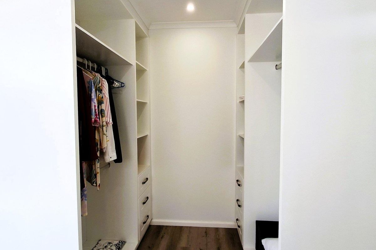 A walk-in wardrobe featuring custom white built-in cabinetry with integrated shelving, drawers, and hanging rails, finished with modern timber-look flooring.