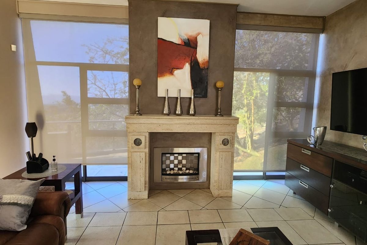 A well-maintained living room featuring a prominent stone fireplace surround, tiled flooring, and large windows with roller blinds providing natural light.