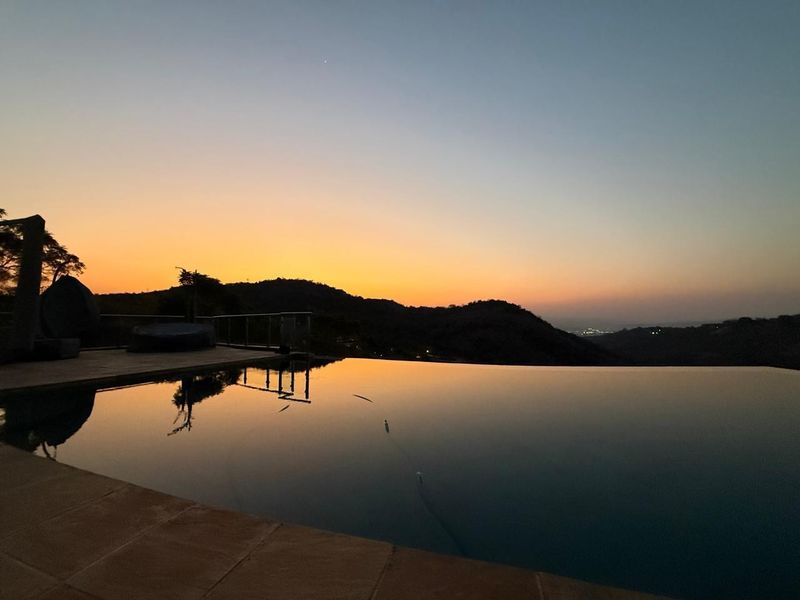 An infinity edge swimming pool overlooking a scenic mountain landscape at sunset, featuring a tiled deck and metal safety railing.