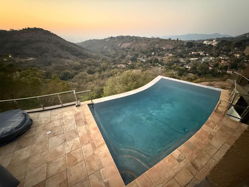An elevated infinity edge swimming pool featuring stone tile decking and a modern glass balustrade, offering expansive panoramic views of the surrounding valley.