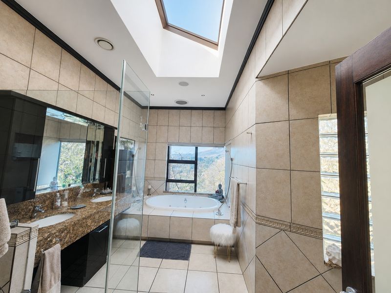 Spacious modern bathroom featuring a large skylight, dual vanity with granite countertops, a built-in bathtub, and a glass-enclosed shower with scenic views.
