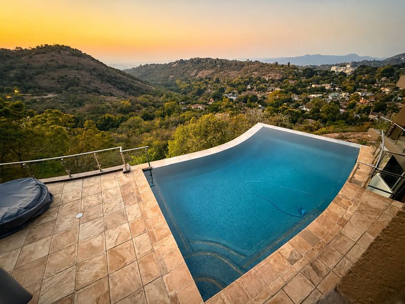 An elevated infinity edge swimming pool featuring stone tile decking and glass balustrades, offering expansive panoramic views of the surrounding valley and landscape.