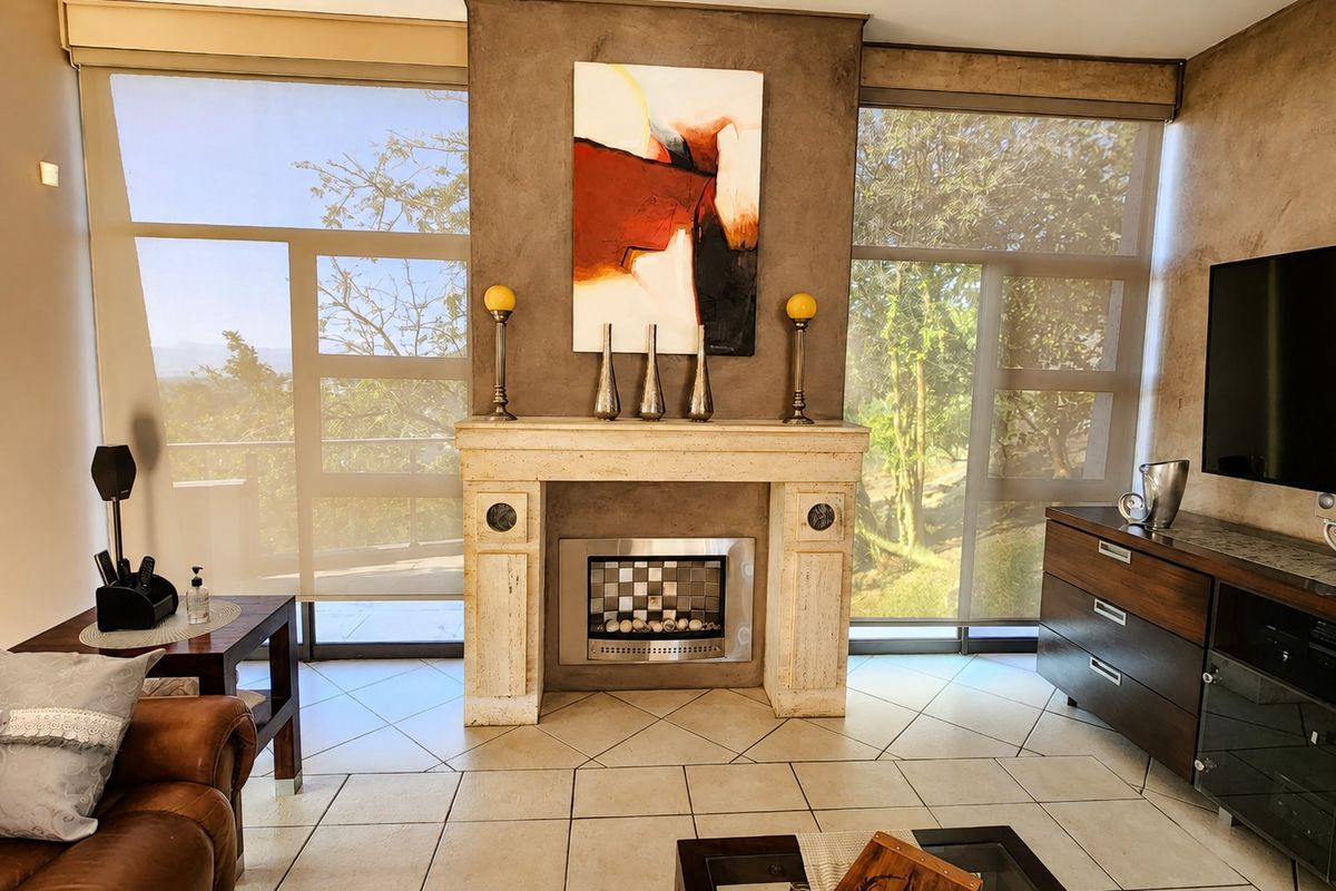 A well-maintained living room featuring a prominent stone fireplace with a built-in gas insert, tiled flooring, and large windows with roller blinds.