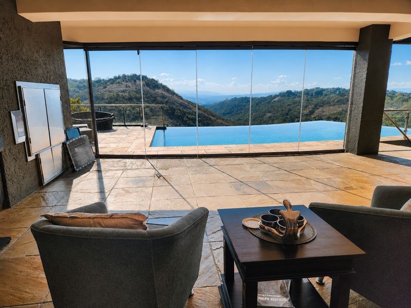Covered patio featuring a built-in braai, stone flooring, and seamless glass stacking doors opening to an infinity pool with panoramic mountain views.
