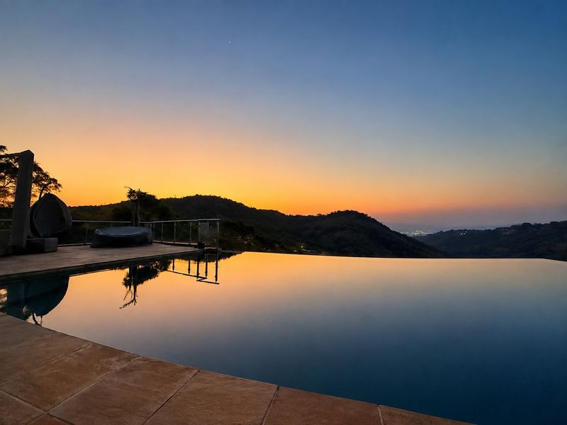 Stunning infinity edge swimming pool featuring a tiled deck and glass balustrade, offering panoramic sunset views over the surrounding hills.