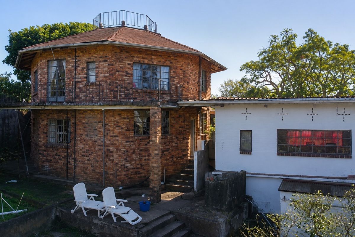 Rear view of a unique two-story circular brick house featuring a roof terrace, balcony, and an attached white-painted structure with concrete stairs.