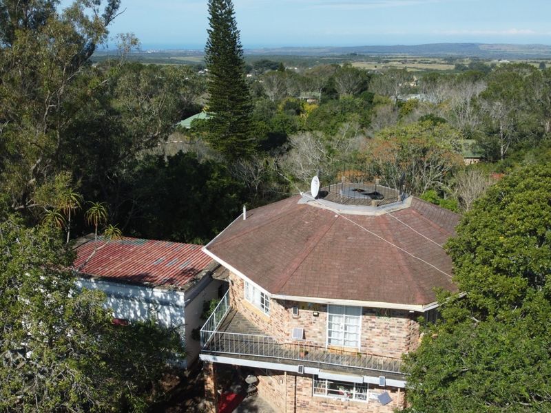 Aerial view of a brick house featuring a tiled roof, balcony with metal railing, and surrounding mature trees in a scenic landscape.
