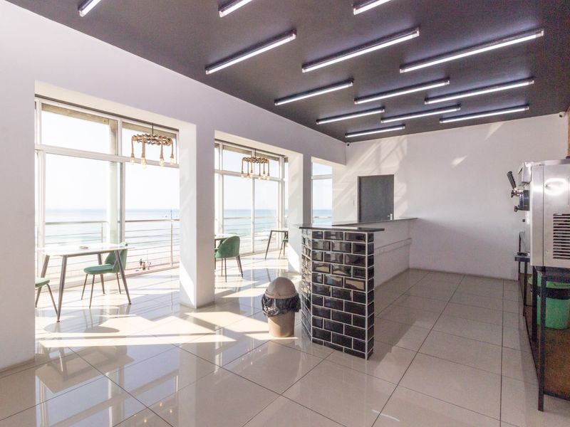 Modern open-plan restaurant space featuring sleek tiled floors, contemporary linear ceiling lighting, and a stylish black subway tile service counter with ocean views.