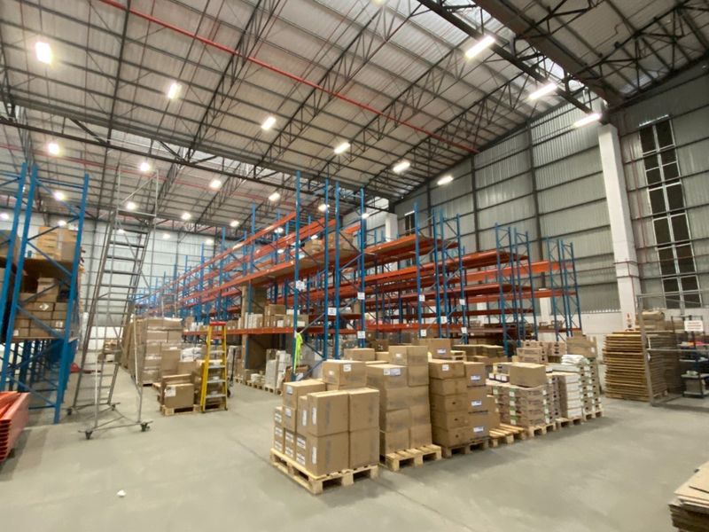 Spacious industrial warehouse featuring high-clearance ceilings, exposed steel roof trusses, high-bay lighting, and extensive heavy-duty steel racking systems on concrete flooring.