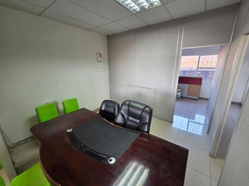 A functional office space featuring suspended ceilings, fluorescent lighting, and tiled flooring, with an adjacent room visible through the doorway.