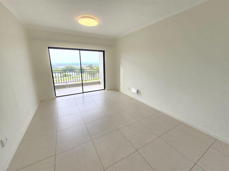 A bright, empty living area featuring neutral tiled flooring and a large sliding glass door that opens onto a balcony with scenic views.