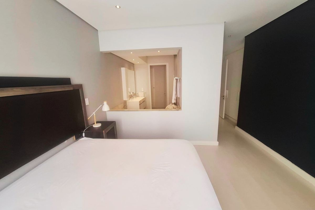 A modern bedroom featuring a semi-open en-suite bathroom layout, recessed lighting, and neutral finishes with a contrasting feature wall.