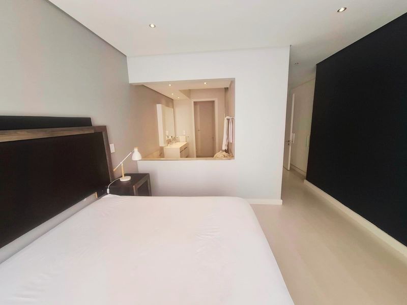 A modern bedroom featuring a semi-open en-suite bathroom layout, recessed lighting, and neutral finishes with a contrasting feature wall.