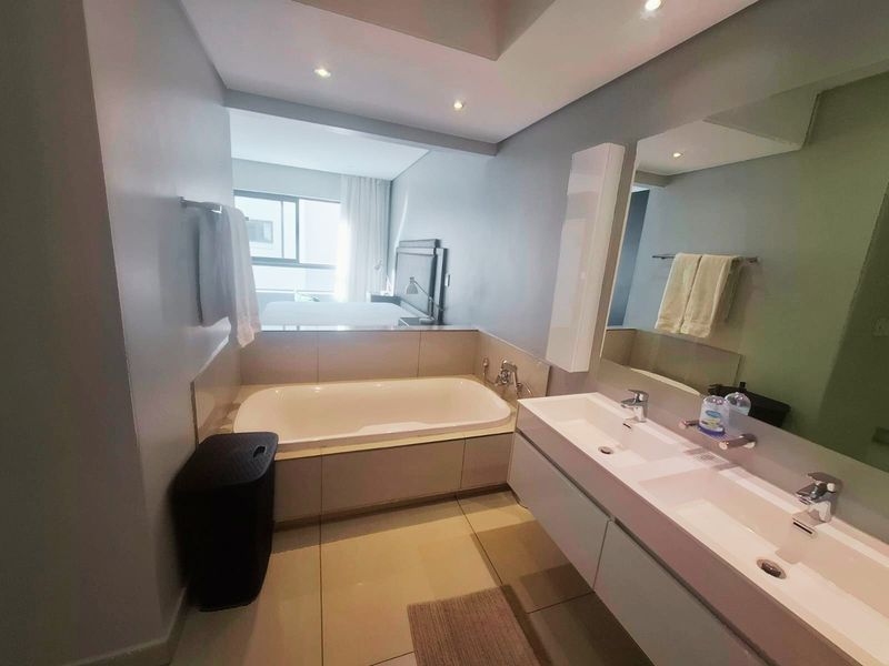 Modern bathroom featuring a built-in bathtub, double vanity with integrated sinks, and large wall-mounted mirror, with an open-plan connection to the bedroom.
