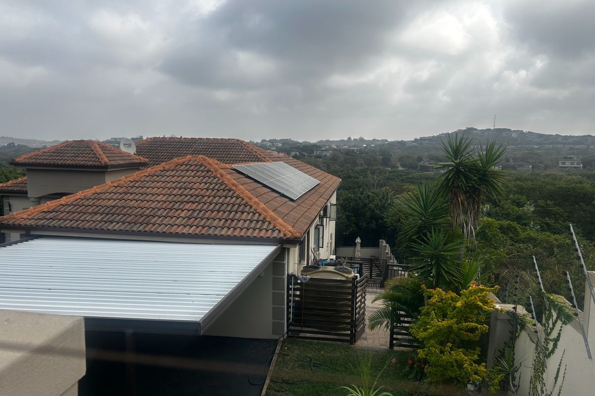 An elevated view of the property featuring a tiled roof with solar panels, a carport, and a water storage tank set within a landscaped garden.