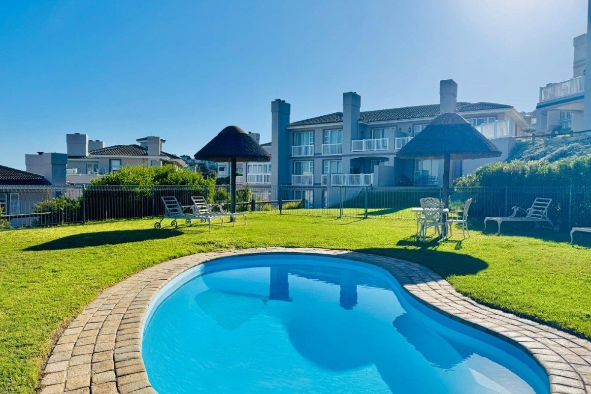 A well-maintained outdoor pool area featuring a kidney-shaped swimming pool with brick coping, surrounded by a manicured lawn and fixed thatched umbrellas.