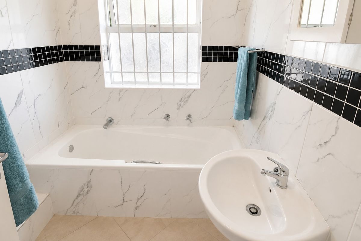 A clean, well-maintained bathroom featuring a built-in bathtub, pedestal washbasin, and marble-look wall tiles with a contrasting black mosaic accent border.