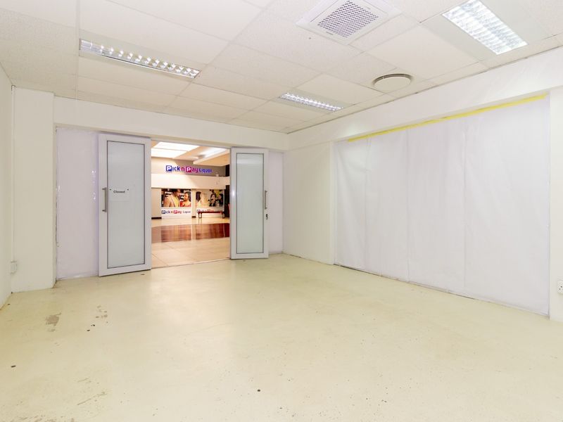 An empty retail utility space featuring a suspended ceiling, recessed lighting, and double glass doors leading to an adjacent commercial area.