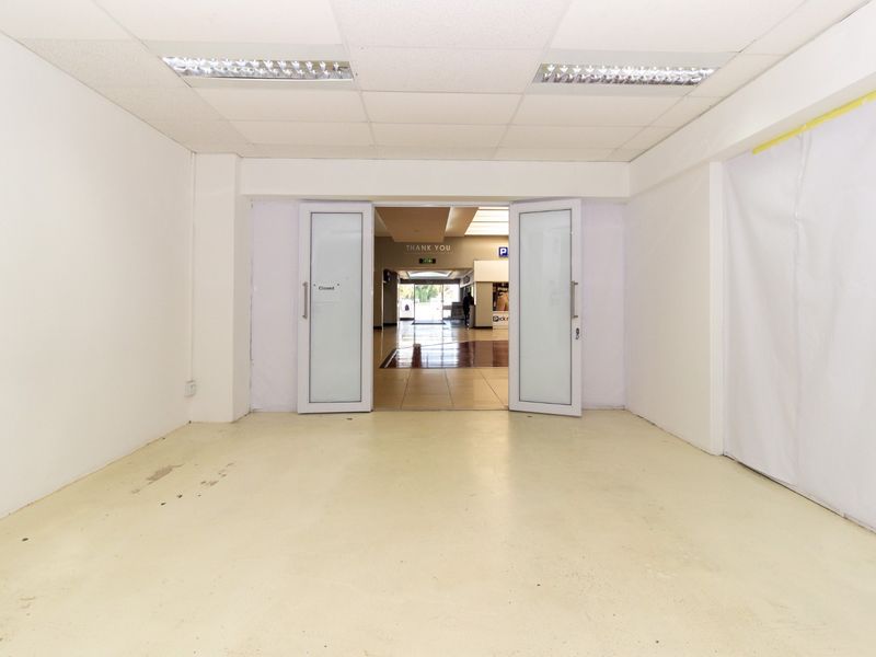 An empty retail unit entrance featuring a suspended ceiling, recessed lighting, and double glass doors leading into a shopping center corridor.
