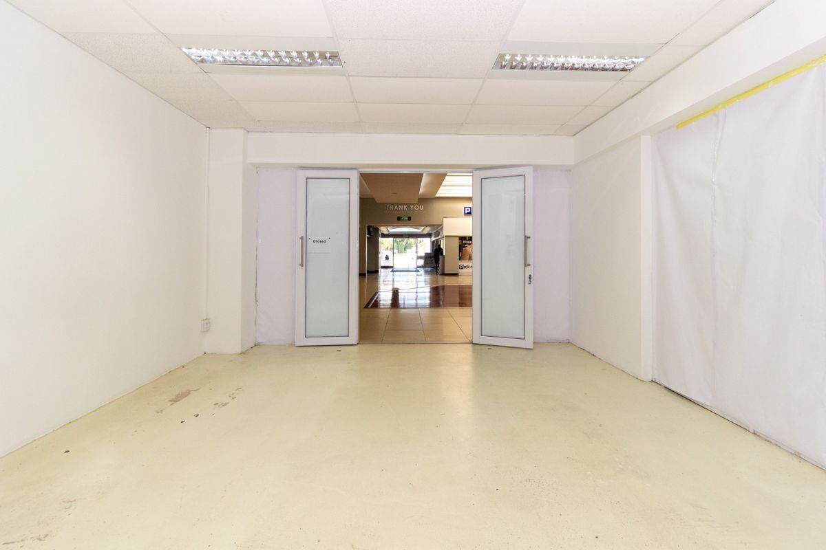 An empty retail unit entrance featuring a suspended ceiling, recessed lighting, and double glass doors leading into a shopping center corridor.