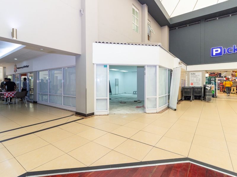 Vacant retail unit located within a shopping center, featuring glass storefront frontage and tiled flooring throughout the common area.