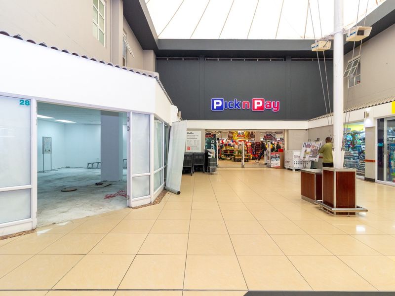 A bright, open-plan retail corridor featuring tiled flooring, a large skylight, and a vacant storefront unit adjacent to a supermarket.