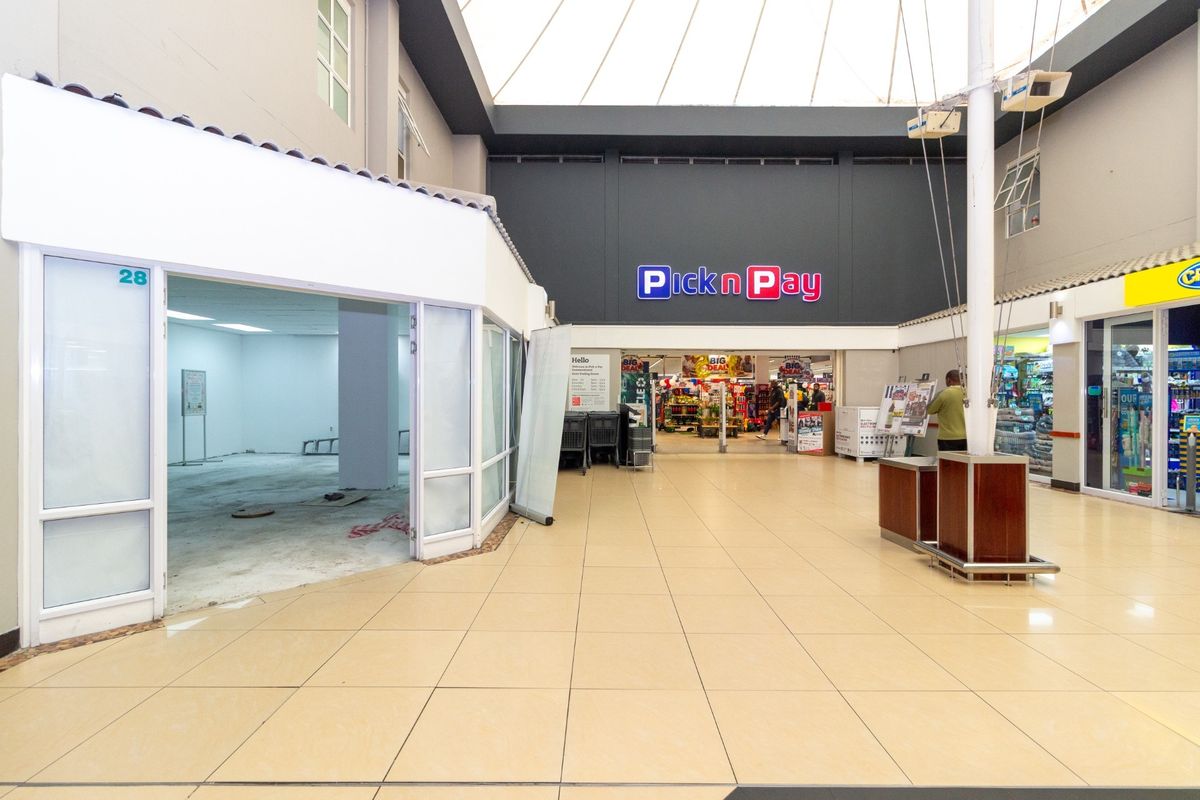 A bright, open-plan retail corridor featuring tiled flooring, a large skylight, and a vacant storefront unit adjacent to a supermarket.
