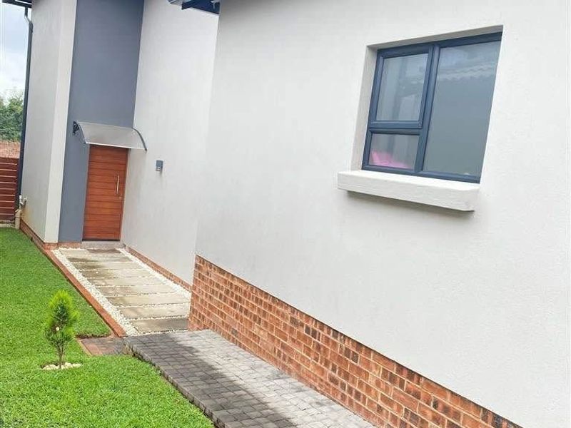 A well-maintained rear exterior view of the townhouse featuring a paved walkway, brick base detailing, and modern aluminium window frames.