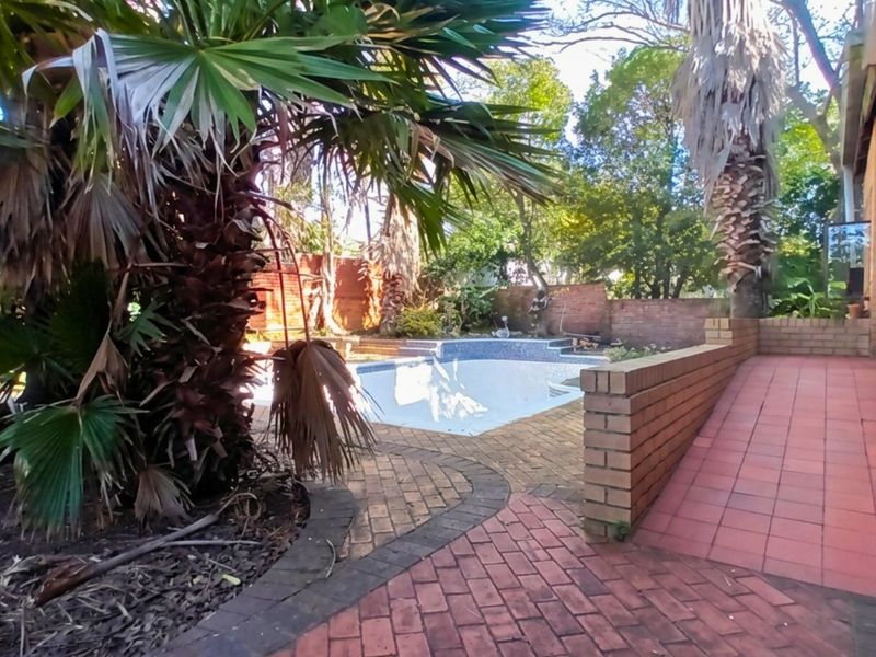Outdoor pool area featuring a swimming pool, brick paving, and a tiled ramp, surrounded by established trees and a brick boundary wall.
