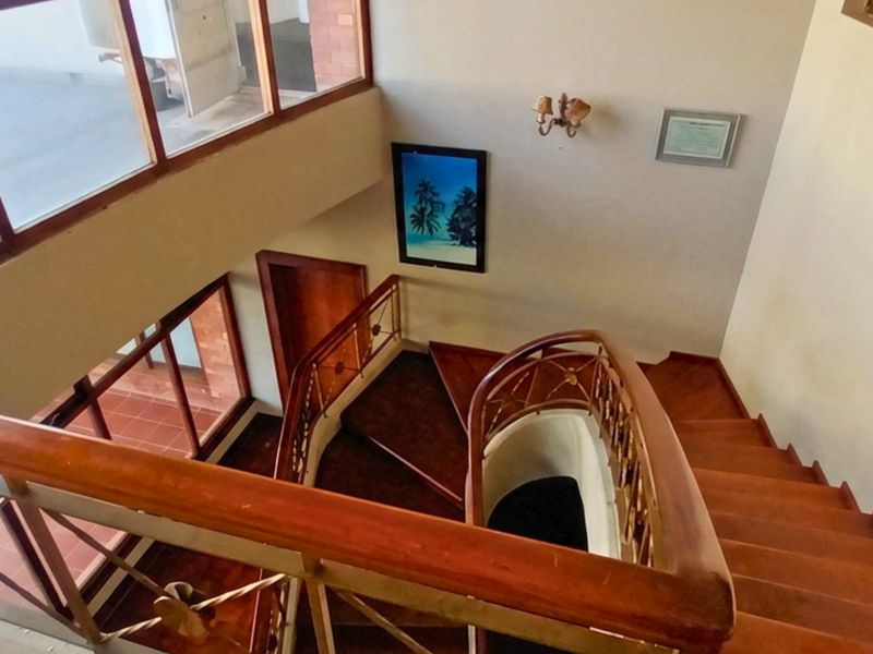 A bright, multi-level hallway featuring a classic wooden staircase with a timber handrail and metal balustrade, illuminated by large windows.