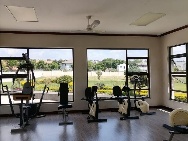 A spacious, well-lit gym area featuring laminate flooring, a ceiling fan, and large windows providing ample natural light and views of the surrounding grounds.