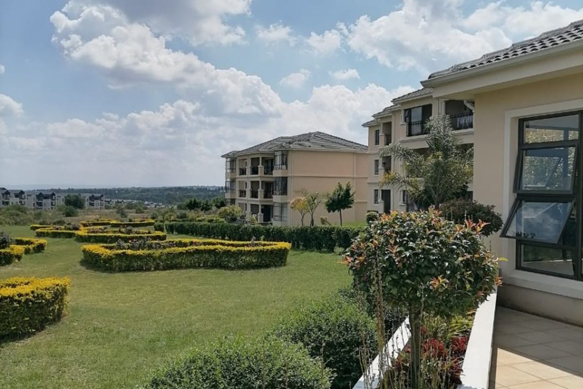 A well-maintained rear exterior view of the apartment complex featuring a tiled patio, landscaped garden beds, and expansive green lawns.
