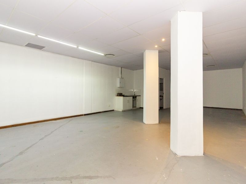An open-plan retail space featuring structural columns, a suspended ceiling, and a small kitchenette area with a sink.