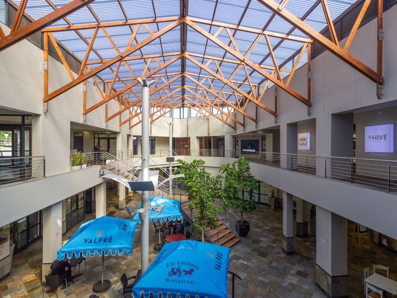 Spacious multi-level retail atrium featuring a high timber truss roof with translucent panels, stone flooring, and mezzanine walkways overlooking a central courtyard area.